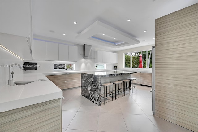 kitchen with a kitchen island, white cabinetry, sink, a kitchen bar, and wall chimney range hood