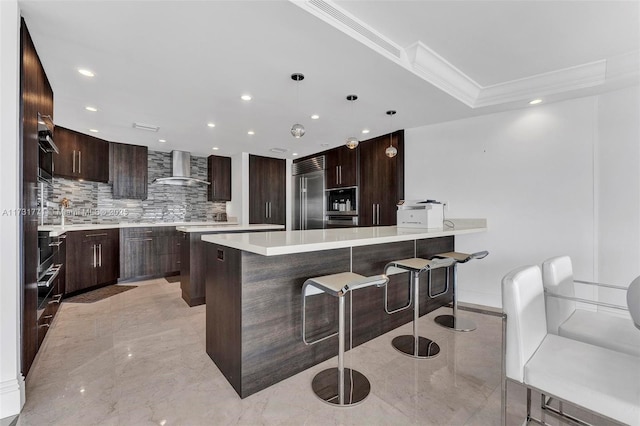 kitchen featuring dark brown cabinetry, crown molding, built in appliances, decorative light fixtures, and wall chimney range hood