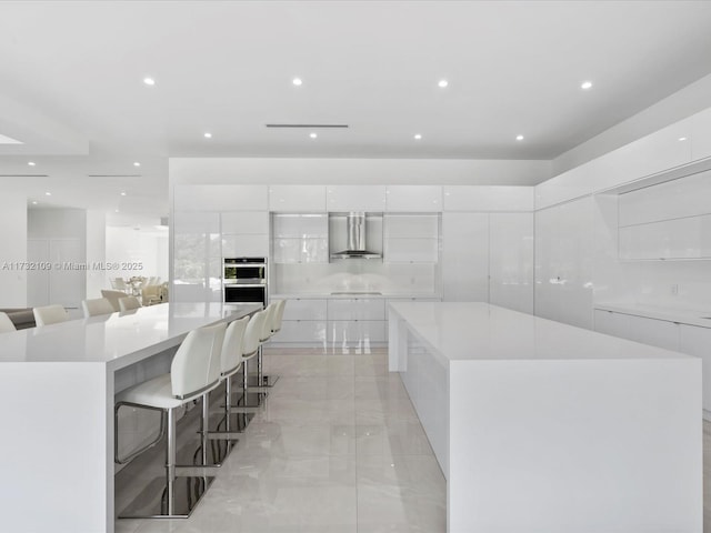 kitchen with a large island, wall chimney exhaust hood, a kitchen breakfast bar, and white cabinets