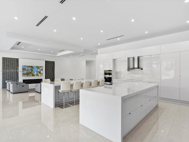 kitchen with a large island, wall chimney range hood, double oven, a tray ceiling, and white cabinets