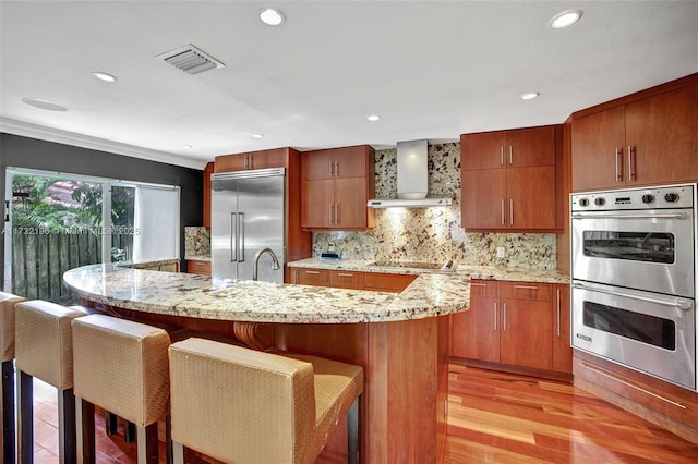 kitchen featuring wall chimney range hood, stainless steel appliances, a kitchen breakfast bar, light hardwood / wood-style floors, and decorative backsplash