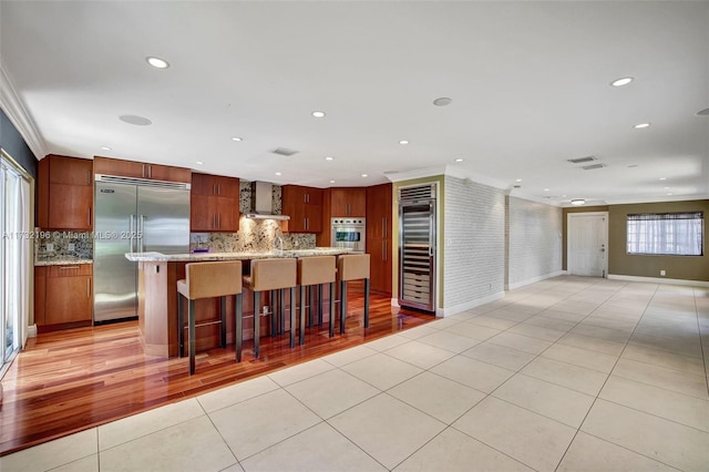 kitchen featuring built in fridge, beverage cooler, a kitchen breakfast bar, ornamental molding, and wall chimney exhaust hood