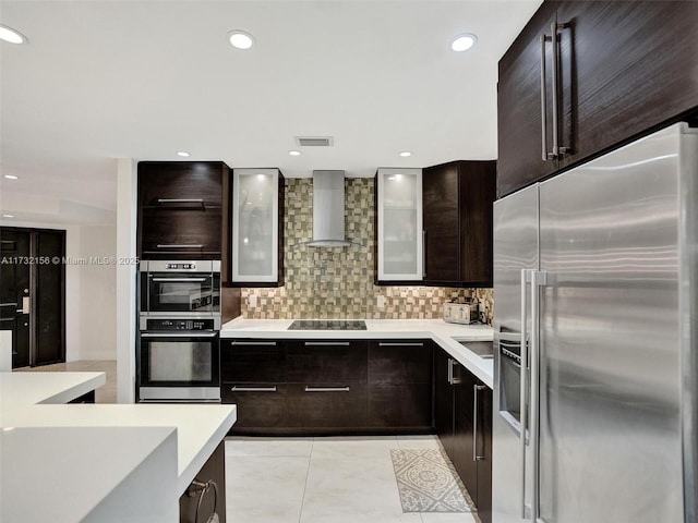 kitchen featuring light tile patterned flooring, dark brown cabinets, wall chimney range hood, stainless steel appliances, and backsplash