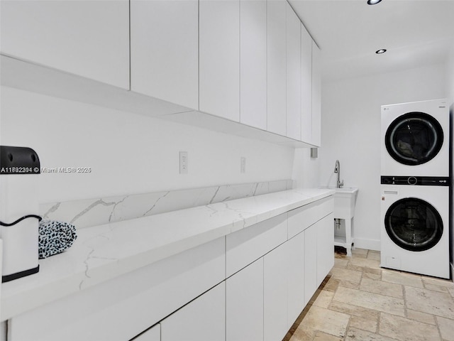 laundry area featuring recessed lighting, stone tile flooring, cabinet space, stacked washer / dryer, and a sink