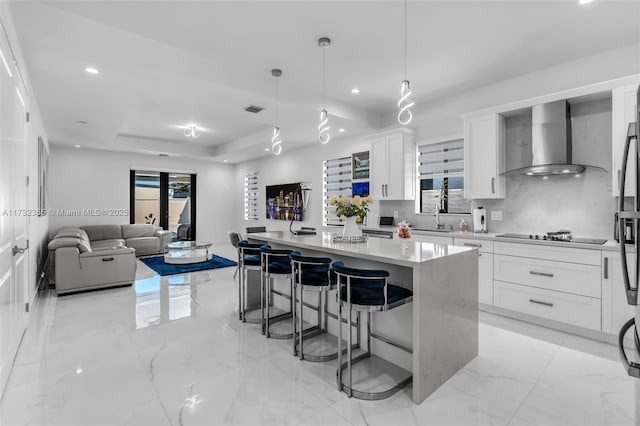 kitchen featuring a center island, hanging light fixtures, a tray ceiling, wall chimney range hood, and white cabinets