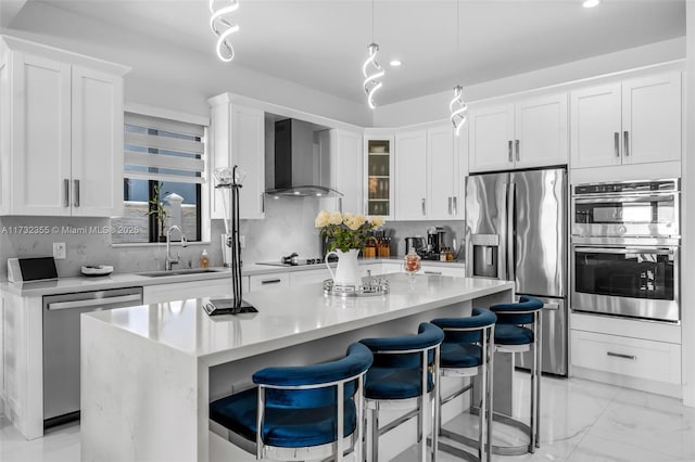 kitchen with wall chimney range hood, sink, appliances with stainless steel finishes, white cabinetry, and an island with sink