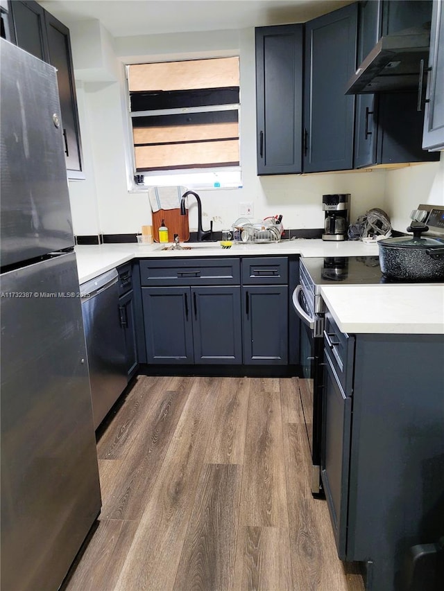 kitchen with light countertops, appliances with stainless steel finishes, a sink, wood finished floors, and under cabinet range hood