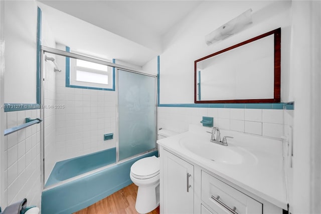 full bathroom featuring bath / shower combo with glass door, tile walls, hardwood / wood-style flooring, vanity, and toilet