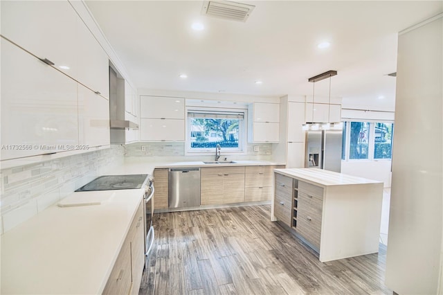 kitchen with wall chimney exhaust hood, sink, hanging light fixtures, appliances with stainless steel finishes, and light hardwood / wood-style floors