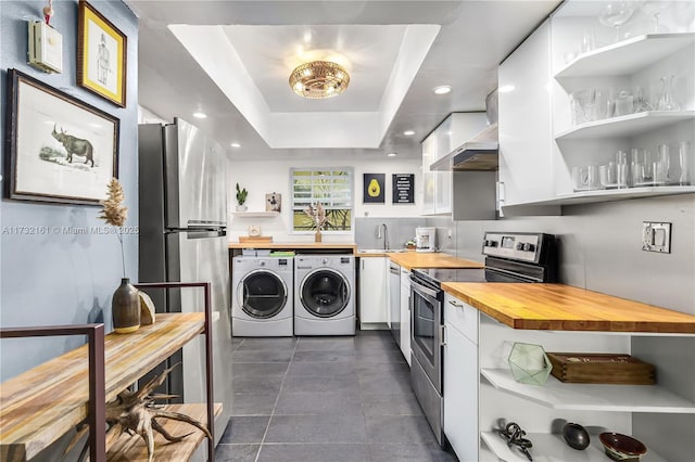 kitchen with butcher block countertops, a tray ceiling, stainless steel appliances, washer and clothes dryer, and wall chimney exhaust hood