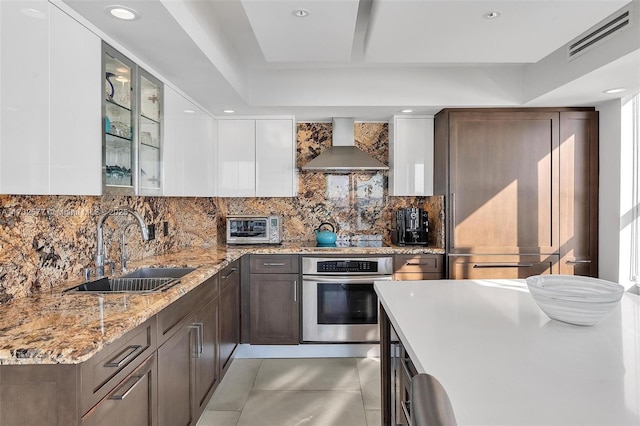 kitchen with sink, light stone counters, white cabinets, oven, and wall chimney exhaust hood