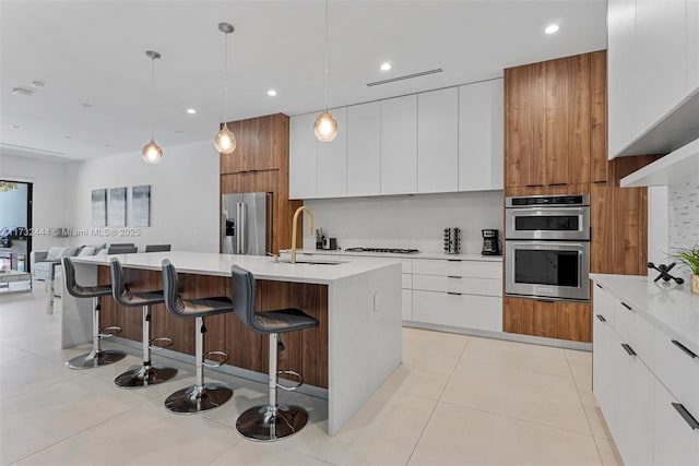 kitchen with appliances with stainless steel finishes, white cabinetry, sink, hanging light fixtures, and a kitchen island with sink