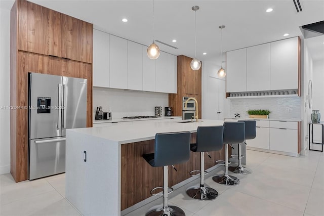 kitchen featuring a breakfast bar area, white cabinetry, decorative light fixtures, stainless steel fridge, and a kitchen island with sink