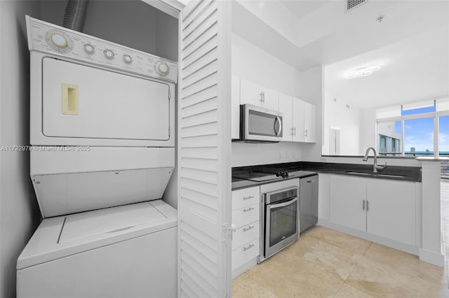 kitchen featuring white cabinets, dark countertops, a sink, stacked washing maching and dryer, and appliances with stainless steel finishes