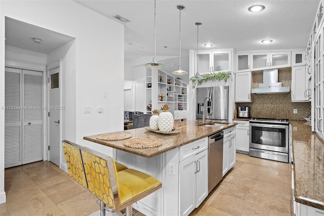 kitchen with wall chimney range hood, a kitchen island with sink, stainless steel appliances, white cabinets, and decorative light fixtures