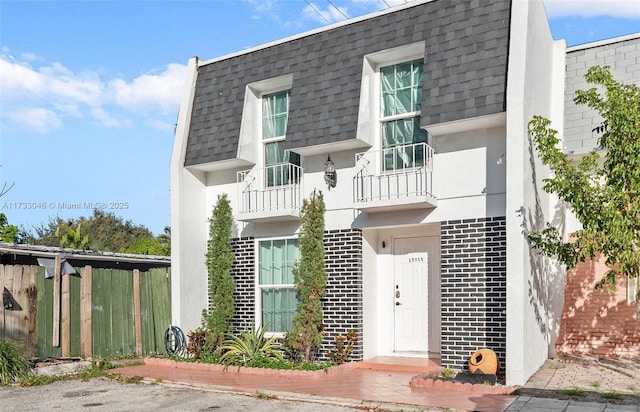 view of front of house featuring mansard roof, brick siding, roof with shingles, and fence