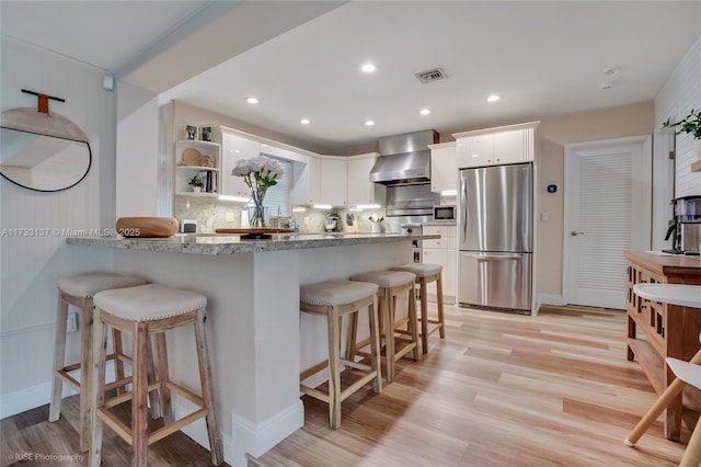 kitchen featuring wall chimney range hood, stainless steel refrigerator, white cabinetry, light stone counters, and a kitchen bar