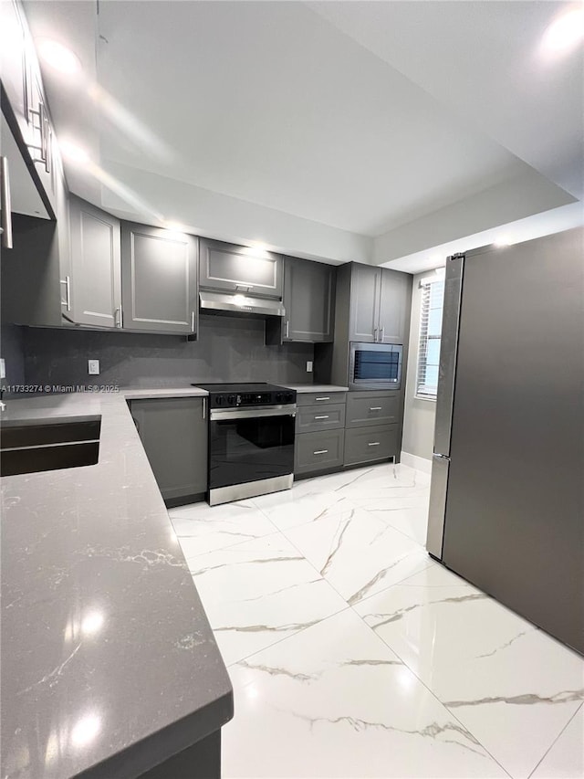 kitchen with a sink, stone counters, gray cabinetry, under cabinet range hood, and appliances with stainless steel finishes