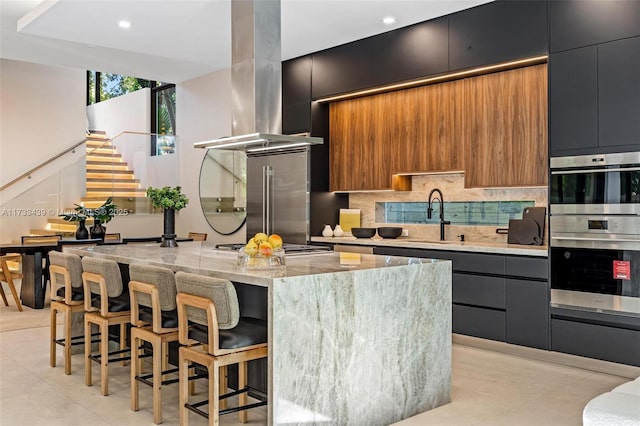 kitchen with island range hood, appliances with stainless steel finishes, a kitchen breakfast bar, light stone countertops, and backsplash