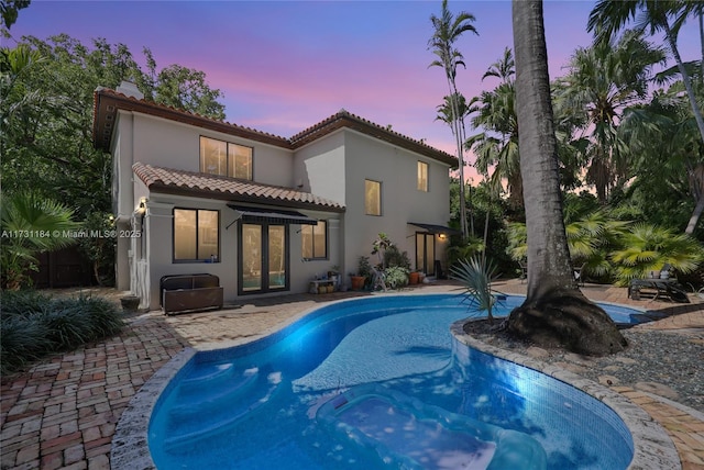 back of house at dusk featuring french doors, a tile roof, a patio, stucco siding, and an outdoor pool