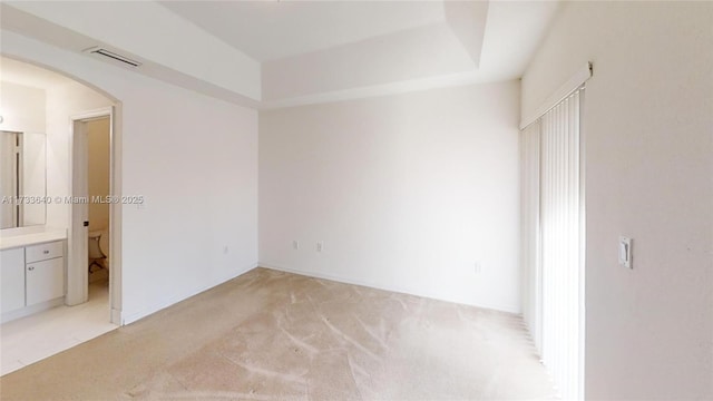 unfurnished bedroom with a raised ceiling, ensuite bathroom, and light carpet