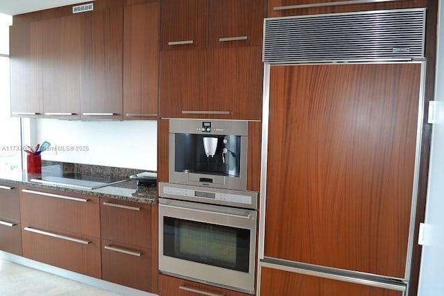 kitchen featuring paneled refrigerator, black electric stovetop, double oven, and dark stone counters