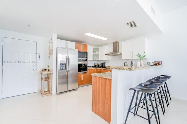 kitchen with wall chimney range hood, stainless steel appliances, white cabinets, a kitchen bar, and kitchen peninsula