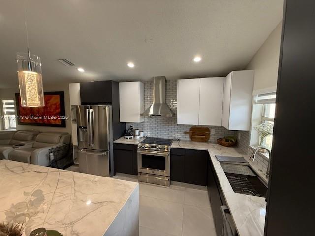 kitchen featuring wall chimney range hood, white cabinetry, backsplash, stainless steel appliances, and decorative light fixtures