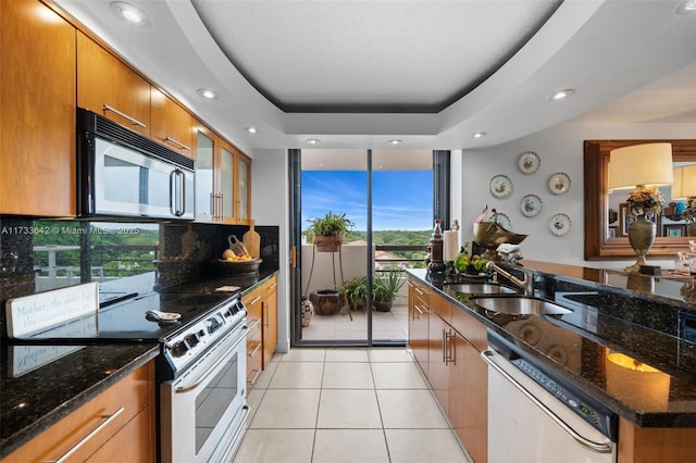 kitchen featuring sink, light tile patterned floors, appliances with stainless steel finishes, a tray ceiling, and dark stone counters