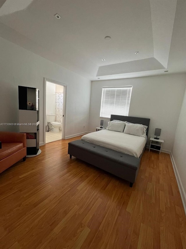 bedroom with ensuite bathroom, a raised ceiling, and light wood-type flooring