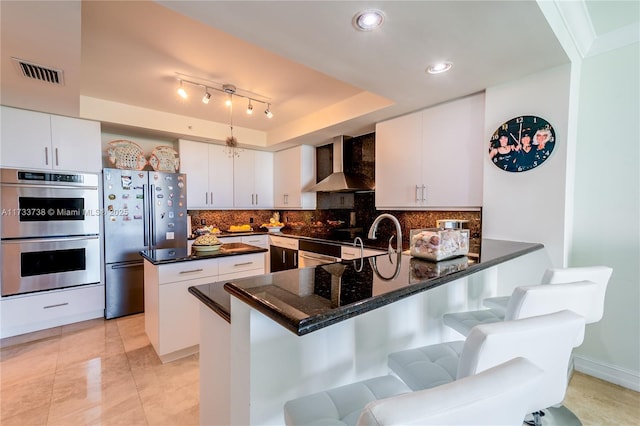 kitchen featuring a breakfast bar, white cabinetry, kitchen peninsula, stainless steel appliances, and wall chimney range hood