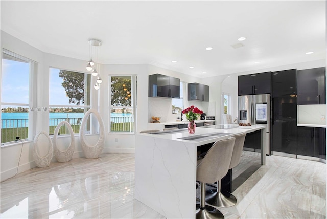 kitchen featuring a kitchen bar, a water view, hanging light fixtures, stainless steel fridge, and wall chimney range hood