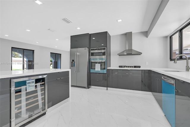 kitchen with wine cooler, gray cabinetry, a sink, appliances with stainless steel finishes, and wall chimney exhaust hood