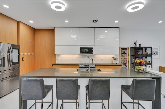 kitchen featuring sink, stainless steel fridge, white cabinetry, a kitchen island with sink, and built in microwave