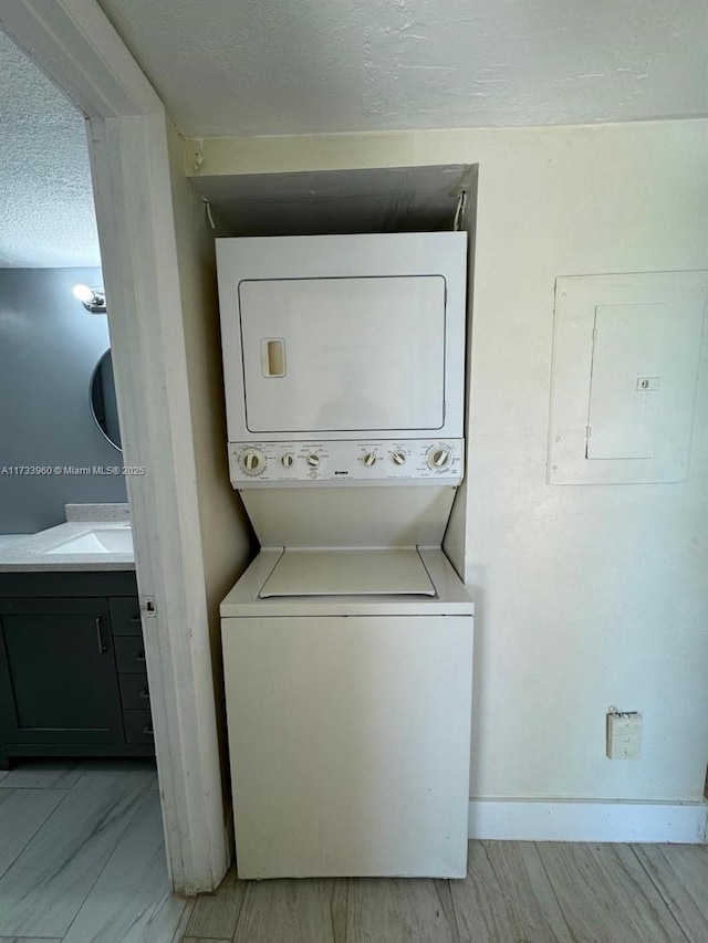 clothes washing area featuring sink, light wood-type flooring, stacked washer and dryer, electric panel, and a textured ceiling