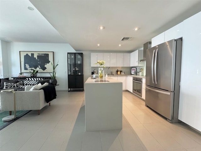 kitchen with white cabinets, a kitchen island with sink, light tile patterned floors, stainless steel appliances, and wall chimney range hood