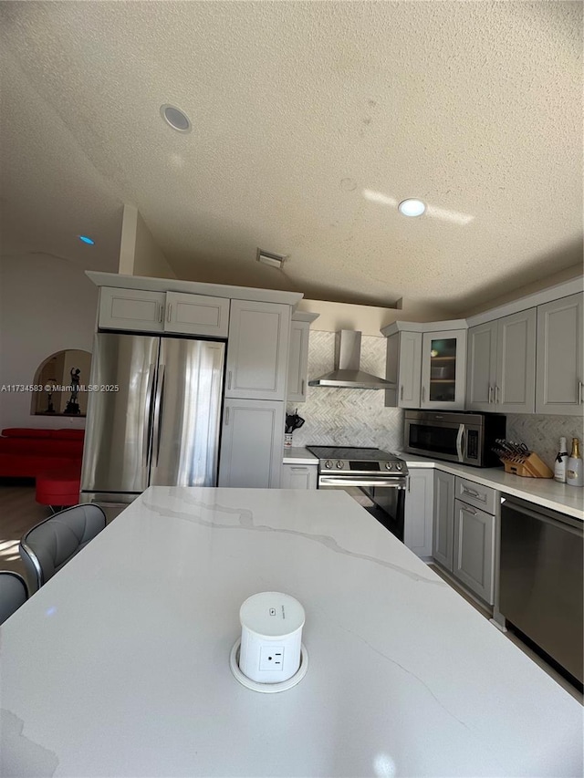 kitchen featuring wall chimney range hood, gray cabinets, appliances with stainless steel finishes, tasteful backsplash, and light stone counters