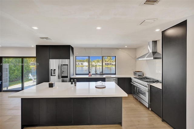 kitchen featuring appliances with stainless steel finishes, an island with sink, sink, wall chimney range hood, and light wood-type flooring