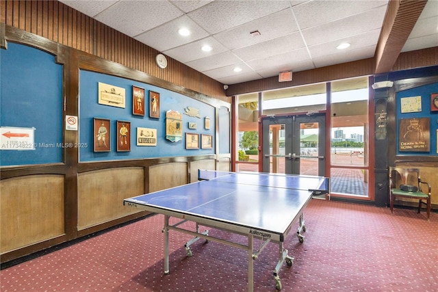 rec room featuring a paneled ceiling, french doors, wooden walls, and carpet flooring