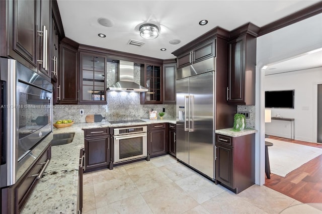 kitchen with light stone counters, built in appliances, dark brown cabinets, decorative backsplash, and wall chimney range hood