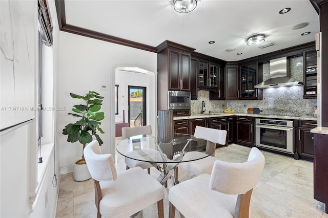 kitchen featuring stainless steel appliances, dark brown cabinets, wall chimney range hood, and backsplash