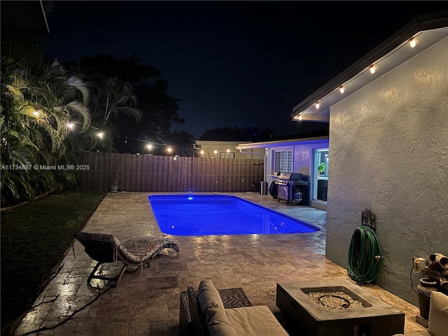 pool at night featuring a grill, a patio area, and a fire pit