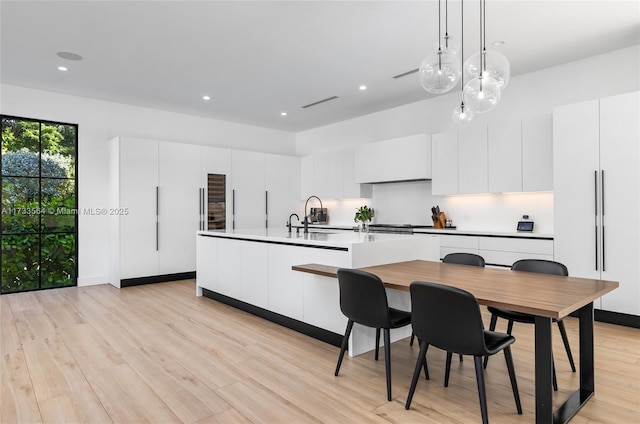 kitchen with light wood-type flooring, decorative light fixtures, a kitchen island with sink, and white cabinets