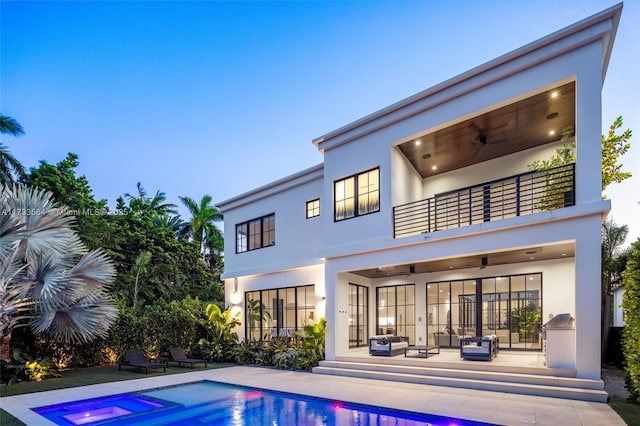 back house at dusk featuring a balcony, a swimming pool with hot tub, and a patio