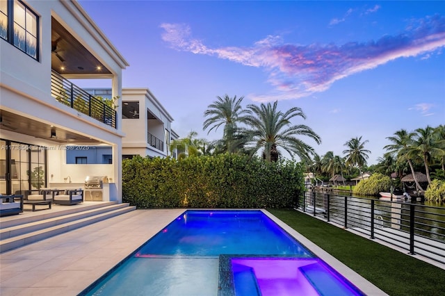 pool at dusk with an outdoor kitchen, a grill, an in ground hot tub, ceiling fan, and a patio area