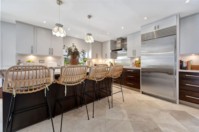 kitchen featuring dark brown cabinetry, a kitchen bar, decorative light fixtures, built in fridge, and wall chimney range hood