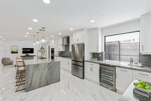 kitchen with appliances with stainless steel finishes, white cabinets, beverage cooler, a kitchen island with sink, and wall chimney range hood