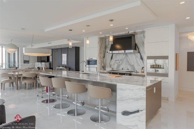 kitchen with white cabinetry, hanging light fixtures, a large island, decorative backsplash, and wall chimney range hood