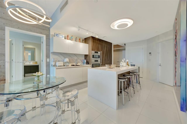 kitchen with light tile patterned floors, white cabinetry, a kitchen island with sink, tasteful backsplash, and decorative light fixtures