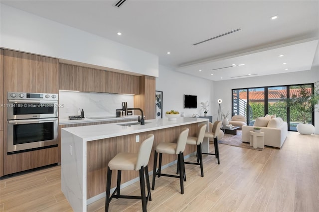 kitchen featuring light hardwood / wood-style flooring, a kitchen island with sink, a kitchen breakfast bar, tasteful backsplash, and stainless steel double oven